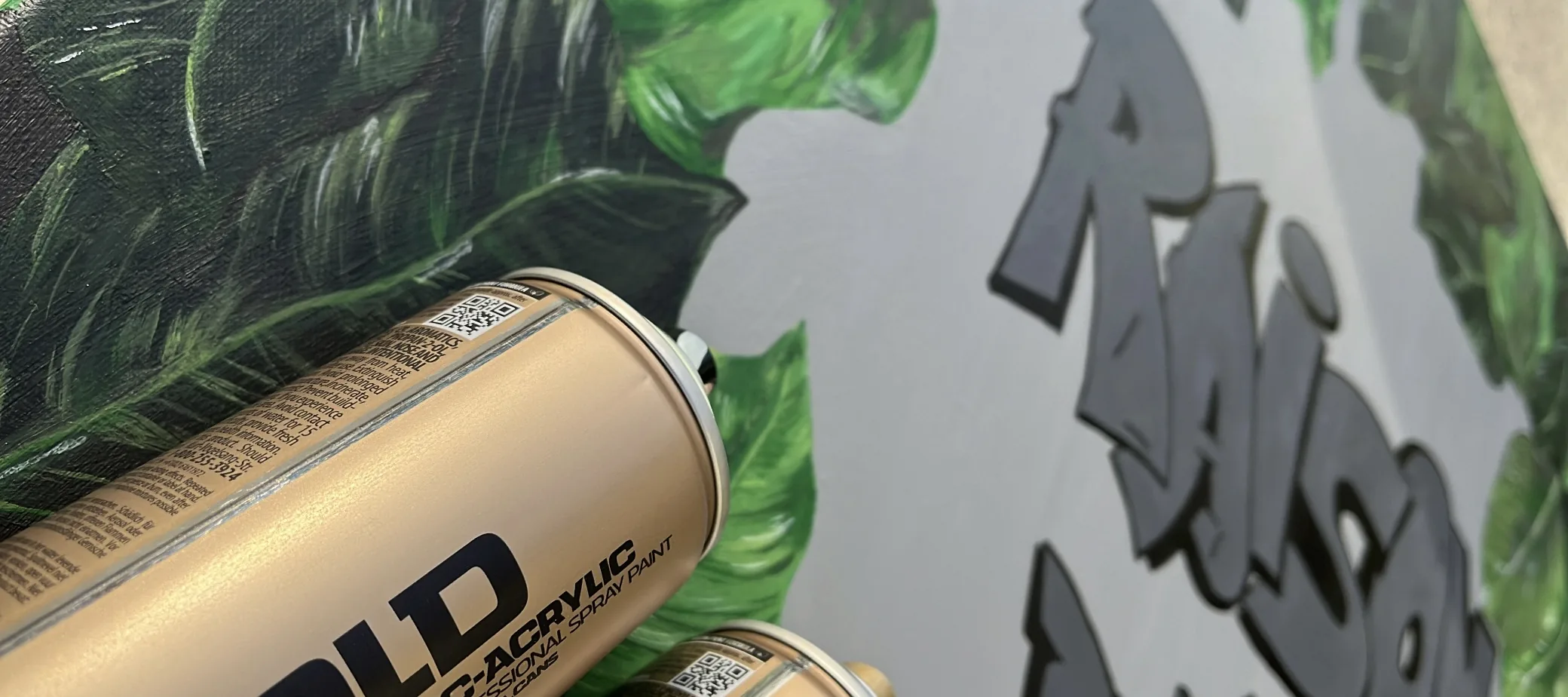 Two gold spray cans lying against “Raison d’être”, its painted leaves and graffiti lettering behind them.