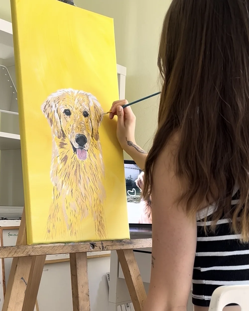 Sylvia at her easel, painting a golden retriever on a yellow canvas.